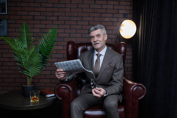 Cheerful, handsome, old, smiling boss in suit reading newspaper,  sitting in leather chair in workstation. - Фото, зображення