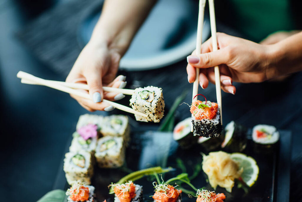 Chopstick holding sushi rolls set with salmon and cream cheese and cuccumber on black plate. Close Up. Uramaki, nori maki or futomaki sushi with trout fillets, soy sauce and wasabi. - Foto, imagen