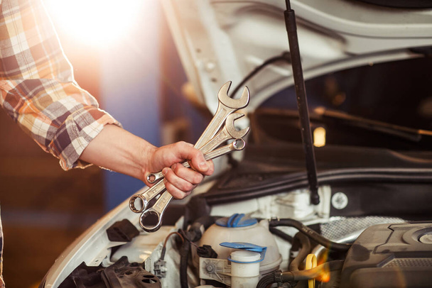 workman hand holding different wrenches with open car hood in garage, sun effect - 写真・画像