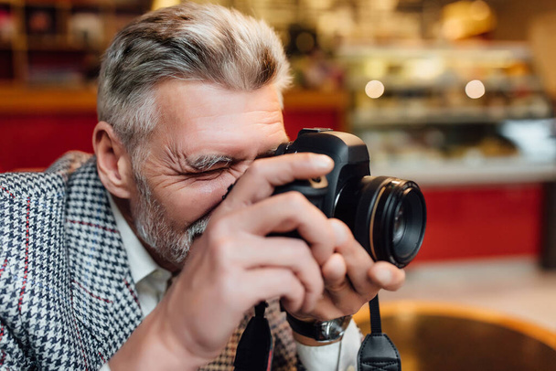 Senior man photographer is holding an old photocamera and make a pictures at modern cafeteria. - Zdjęcie, obraz