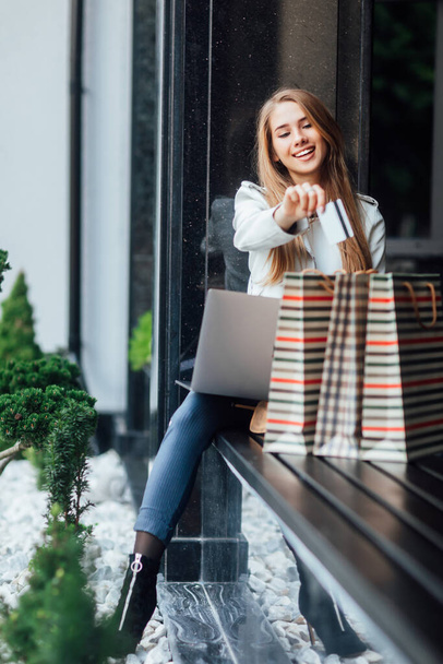 Blonde girl after buying new clothes, shopaholic sit near mall and doing online shopping with credit card. - Foto, afbeelding