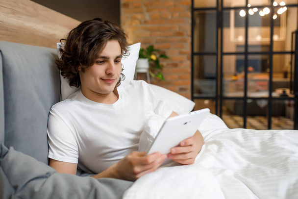 Photo of man laying on his bed at morning time, holding a laptop while reading news. - Foto, Bild