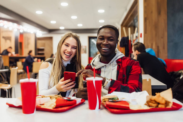 Young,pretty couple spend time at fast food cafe, use smart phone, eating food and smiling. - Фото, изображение