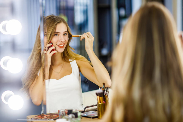 Young beautiful girl doing makeup in front of mirror and talking on mobile phone. - Photo, image