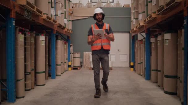 Een jonge mannelijke werknemer bijhouden van records met behulp van digitale tablet wakker door het grote magazijn met vracht gestapeld - Video