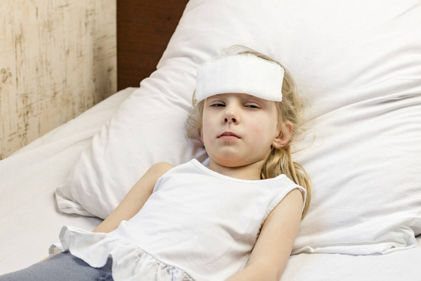 白い髪の少女がベッドに横になっている。彼女は痛みを和らげるために額に包帯をしている。接近中だ。そこには小さな. - 写真・画像
