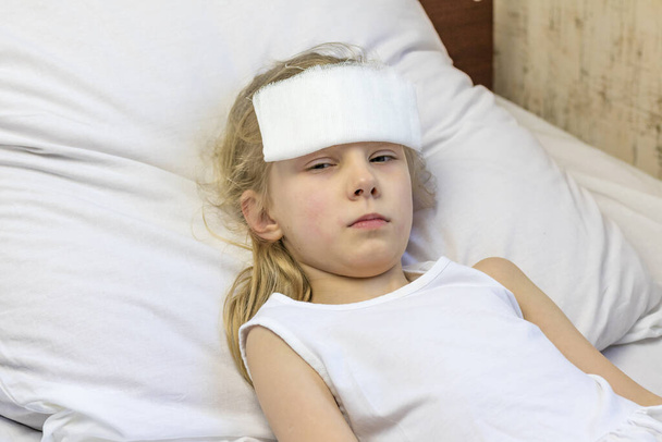 白い髪の少女がベッドに横になっている。彼女は痛みを和らげるために額に包帯をしている。接近中だ。そこには小さな. - 写真・画像