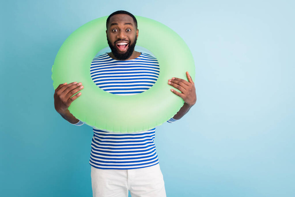 Photo of cheerful dark skin guy hold green life buoy around neck ready swim ocean sea traveler good mood sunny day wear striped sailor shirt isolated blue color background - Foto, immagini