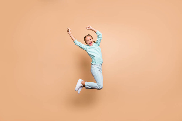 Full body profile photo of beautiful funny little lady jumping high up cheerleader celebrating winning raise fists wear blue sweater jeans footwear isolated beige color background - Foto, imagen