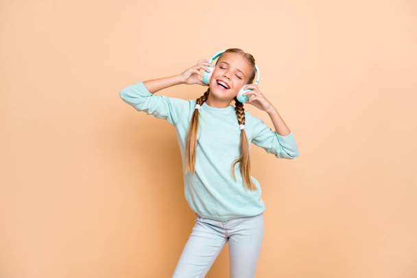 Photo of beautiful funny little lady eyes closed enjoy listen cool wireless earphones favorite song wear blue pullover jeans isolated beige pastel color background - Foto, imagen