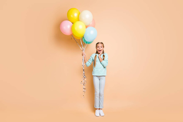 Full length photo of beautiful small lady many colorful air balloons surprise festive birthday party hand on cheek wear blue sweater jeans sneakers isolated beige pastel color background - Φωτογραφία, εικόνα