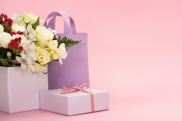 bouquet of flowers in festive gift box with bow near violet paper bag on pink background - Photo, image