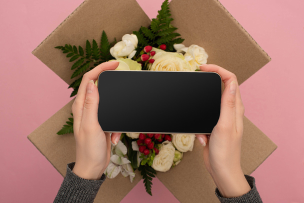 cropped view of woman holding smartphone above bouquet of flowers in cardboard box on pink background - Foto, immagini
