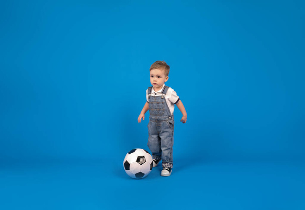 Little football champion. Adorable european baby boy with soccer ball sitting on floor over blue background in studio, copy space, sport concept - Foto, immagini