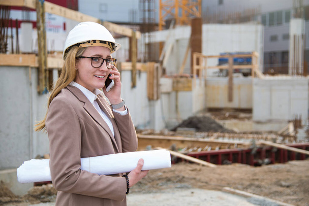 Beautiful young Woman as an architect, engineer with helmet, blueprint, plans on a building site. the architect is calling on a cell phone, mobile. Copy space and construction site with crane in the background - Foto, Bild