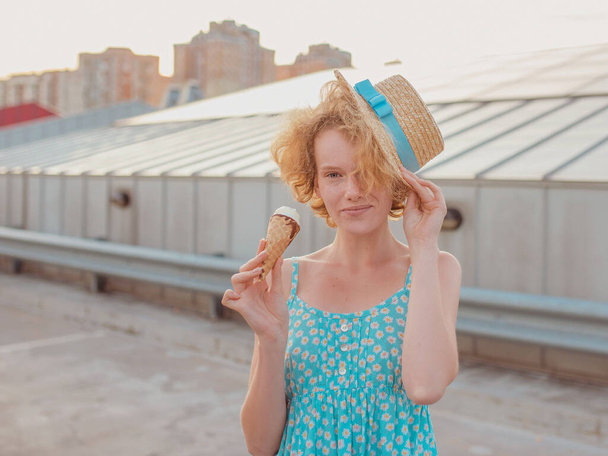 young happy cheerful curly redhead woman in straw hat, blue sundress eating ice cream on skyscraper roof (skyroof). Fun, lifestyle, urban, modern, roof, city, summer, fashion, youth concept - Photo, Image