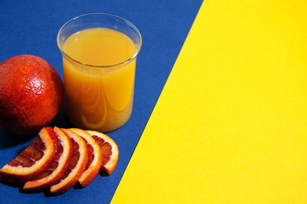 A whole red orange lies on a blue / yellow background. Slices of orange lie nearby and there is a glass of orange juice. Soft focus, top view. Trend blue background - Photo, Image