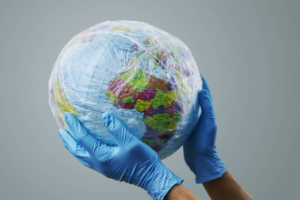 a doctor man wearing blue surgical gloves holding a world globe wrapped in plastic, depicting the plastic contamination or the protection against the epidemic infectious diseases or the air pollution - Photo, image