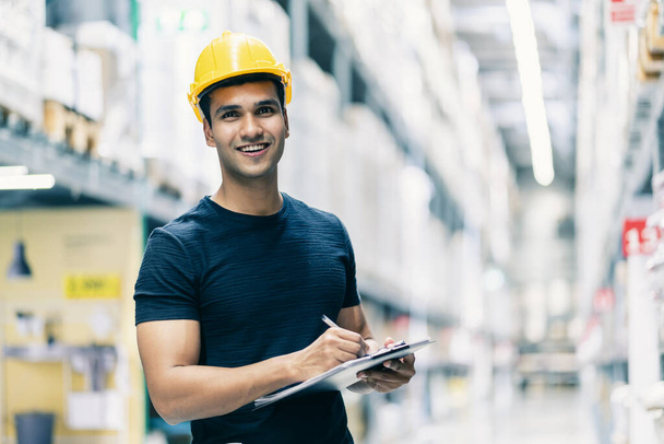 Smart Indian engineer man wearing safety helmet doing stock tick check and cardboard stock product management in factory warehouse background - Фото, зображення