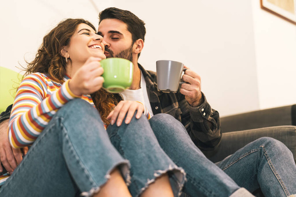 2人の若いカップルがコーヒーカップを隣のソファに座って飲む – 自宅で一緒に優しい瞬間を持っている幸せな恋人 – 愛の関係と若者のライフスタイルの概念 - 写真・画像