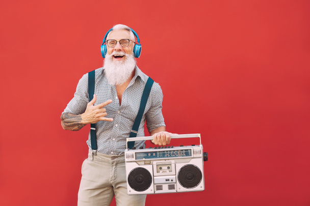 Senior crazy man listening music with headphones and vintage boombox outdoor - Hipster male having fun life in past time - Senioren lifestyle activity - Roter Hintergrund - Foto, Bild