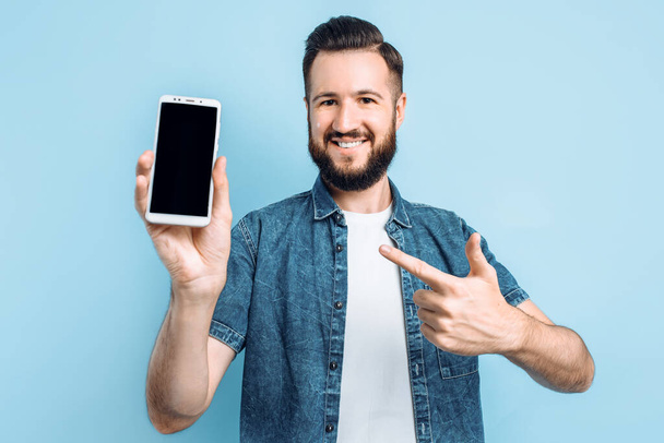 Happy young man holding a smartphone in his hand and showing a blank smartphone screen to the camera, pointing at it with his finger, on a blue bac - Foto, Bild