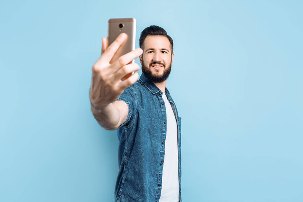 Happy fashionable man with a beard in casual clothes, smiling and taking a selfie on a smartphone, stands on a blue background - Фото, изображение