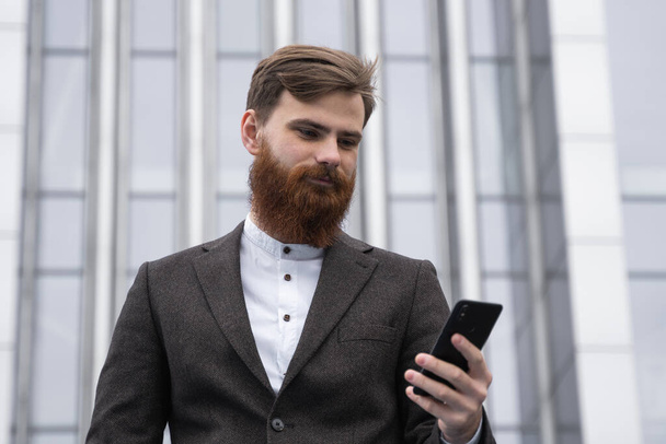Joven barbudo Empresario sosteniendo teléfono inteligente móvil utilizando mensajes de texto SMS aplicación usando chaqueta al aire libre. Empresario exitoso vestido con ropa formal que se comunica con un socio de negocios en línea
. - Foto, imagen