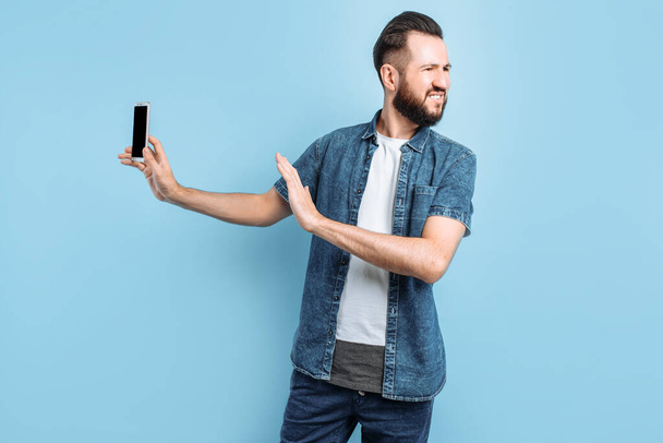 An irritated young man keeps his smartphone away from his ear. An angry person does not want to listen to a conversation on the phone, standing on an isolated blue background - Photo, Image