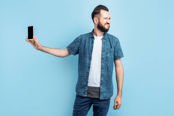 An irritated young man keeps his smartphone away from his ear. An angry person does not want to listen to a conversation on the phone, standing on an isolated blue background - Foto, afbeelding