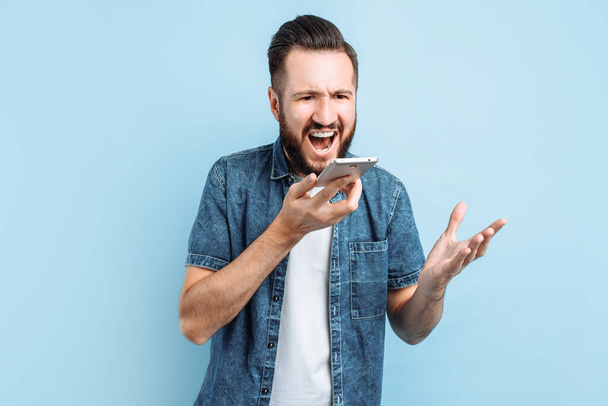 An excitedly shouting man is talking on a smartphone and shouting into the phone, standing on a blue background - Fotoğraf, Görsel