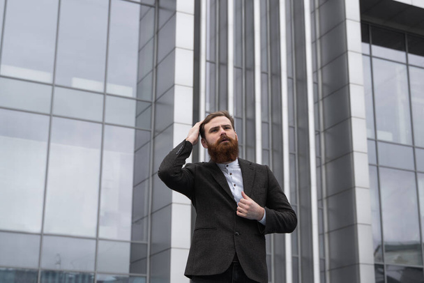 Portrait of an handsome businessman wearing jacket over office building in financial district outdoors. Young bearded Businessman outdoor. Successful entrepreneur dressed in formal wear. - Foto, imagen