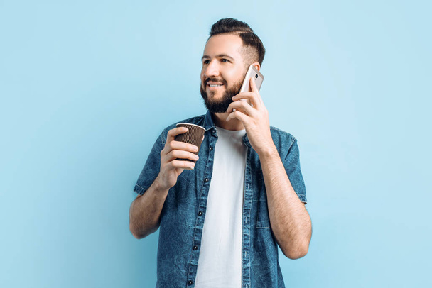 Attractive happy young man with a beard , talking on the phone and drinking coffee or tea, in a paper Cup standing on an isolated blue background - Fotoğraf, Görsel