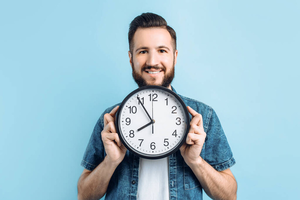 Beautiful stylish happy young man with beard holding round wall clock isolated on blue background - Foto, Bild