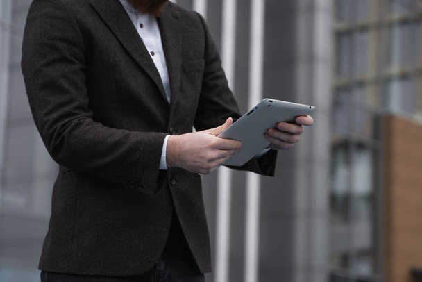 Modern Young bearded Business man working with a digital tablet. Young hipster businessman holding tablet in hands outdoor. Working online with a tablet while standing outside on an office building. - Fotografie, Obrázek