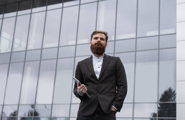 Modern Young bearded Business man standing with a digital tablet. Young hipster businessman holding tablet in hands outdoor. Working online with a tablet while standing outside on an office building. - Valokuva, kuva