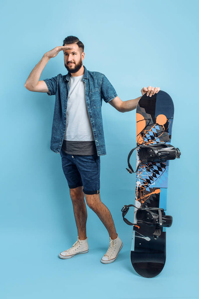 A happy bearded man, dressed in shorts and a shirt , holds a snowboard and looks into the distance, standing on an isolated blue background - Фото, зображення