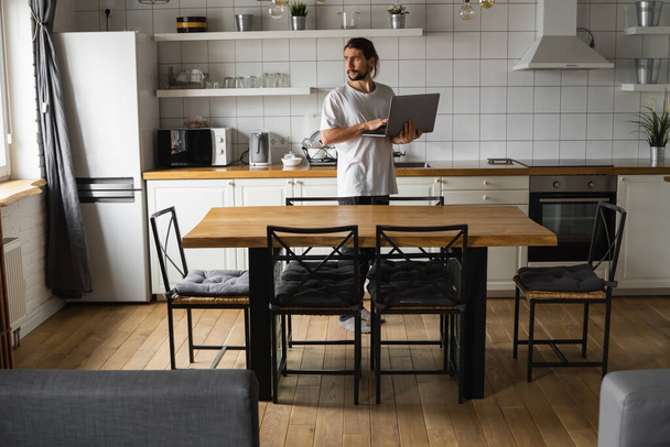 Freelancer working from home on the kitchen and using laptop. Bearded man working with a laptop and reading good news. Handsome successful self entrepreneur standing with laptop and working at his - Valokuva, kuva