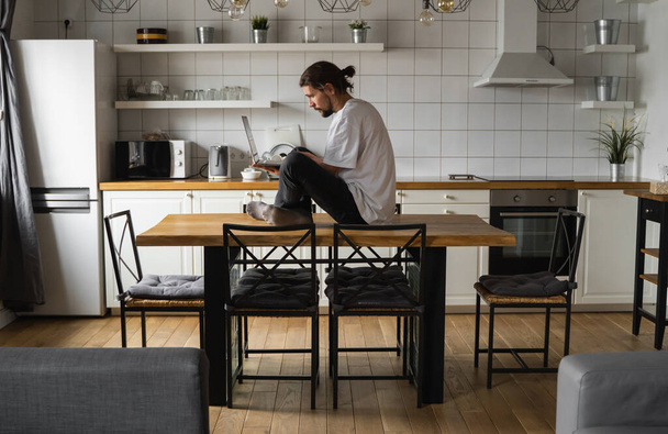Freelancer working from home sitting on a top of the kitchen table and using laptop. Bearded man working with a laptop and reading news. Handsome successful self entrepreneur working at his modern - Фото, изображение