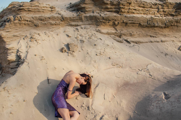 Mulher de cabelos castanhos em roxo brilhante vestido encontra-se na encosta de areia
 - Foto, Imagem