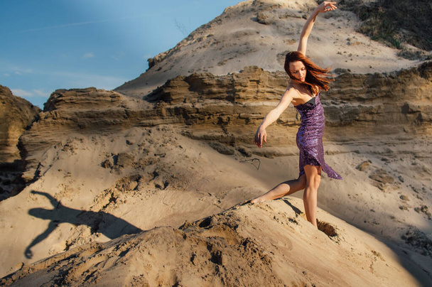 Καστανή γυναίκα με μωβ αστραφτερό φόρεμα που χορεύει στην πλαγιά της άμμου - Φωτογραφία, εικόνα