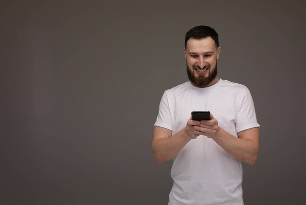 Handsome bearded model is posing with phone in hands over grey background. - Φωτογραφία, εικόνα