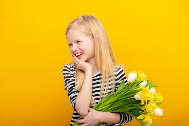 Dreamy smiling little girl with blonde hair holds spring bouquet white tulip and daffodil flowers on yellow studio background. Looks to the side. Spring Woman day. - Fotoğraf, Görsel
