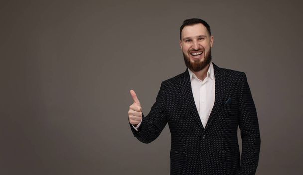 Portrait young smiling bearded businessman. Caucasian guy business suit studio gray background. Modern business person showing thumbs up sign. - Φωτογραφία, εικόνα