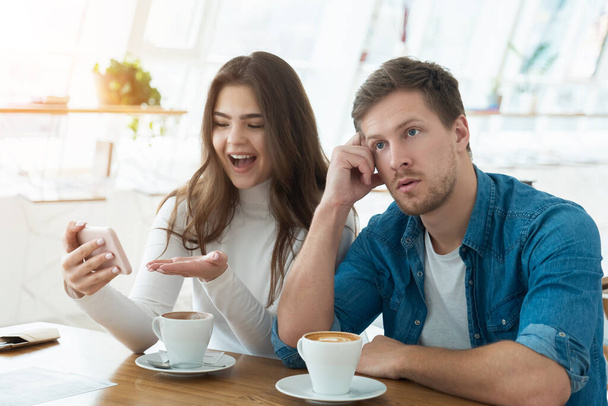 young couple beautiful woman communicates via smartphone and handsome man looks annoyed while drinking coffee in cafe , relationships concept. - 写真・画像