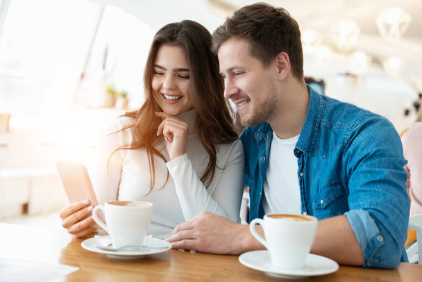 young woman drinks coffee with her husband, she shows him photoes on the phone, both look happy, multitasking concept. - Foto, imagen