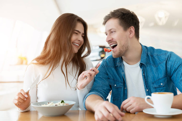 young couple beautiful woman feeds her smiling handsome man with salad drinking coffee in cafe looking happy, relationship concept. - Фото, изображение