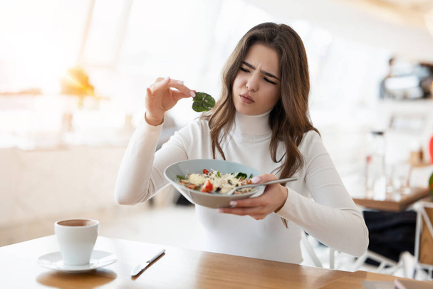 young beautiful woman looks unsatisfied with quality of salad holding piece of green leaf, looking unhappy during lunch time at cafe, , complaining guest concept. - Foto, afbeelding