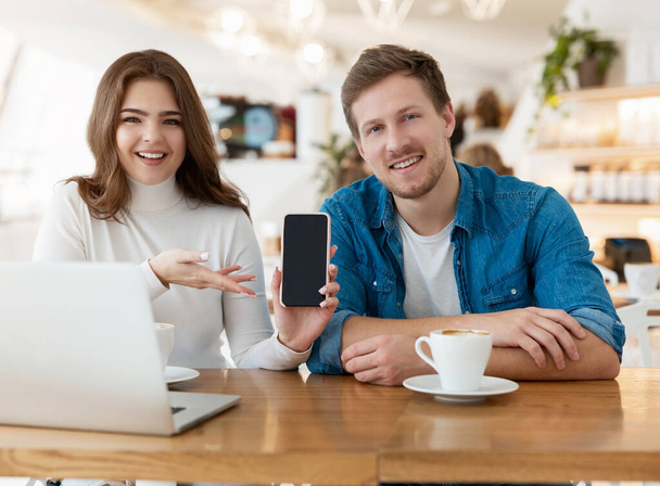 young couple beautiful woman and handsome man sitting at cafe during coffee break, she demonstrates new smartphone device, technology development concept. - 写真・画像