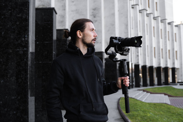 Νέοι επαγγελματίες videographer κρατώντας επαγγελματική φωτογραφική μηχανή σε 3-άξονα gimbal σταθεροποιητή. Pro εξοπλισμός βοηθά να κάνει υψηλής ποιότητας βίντεο χωρίς ανακίνηση. Καμεραμάν με μαύρη κουκούλα που φτιάχνει βίντεο. - Φωτογραφία, εικόνα